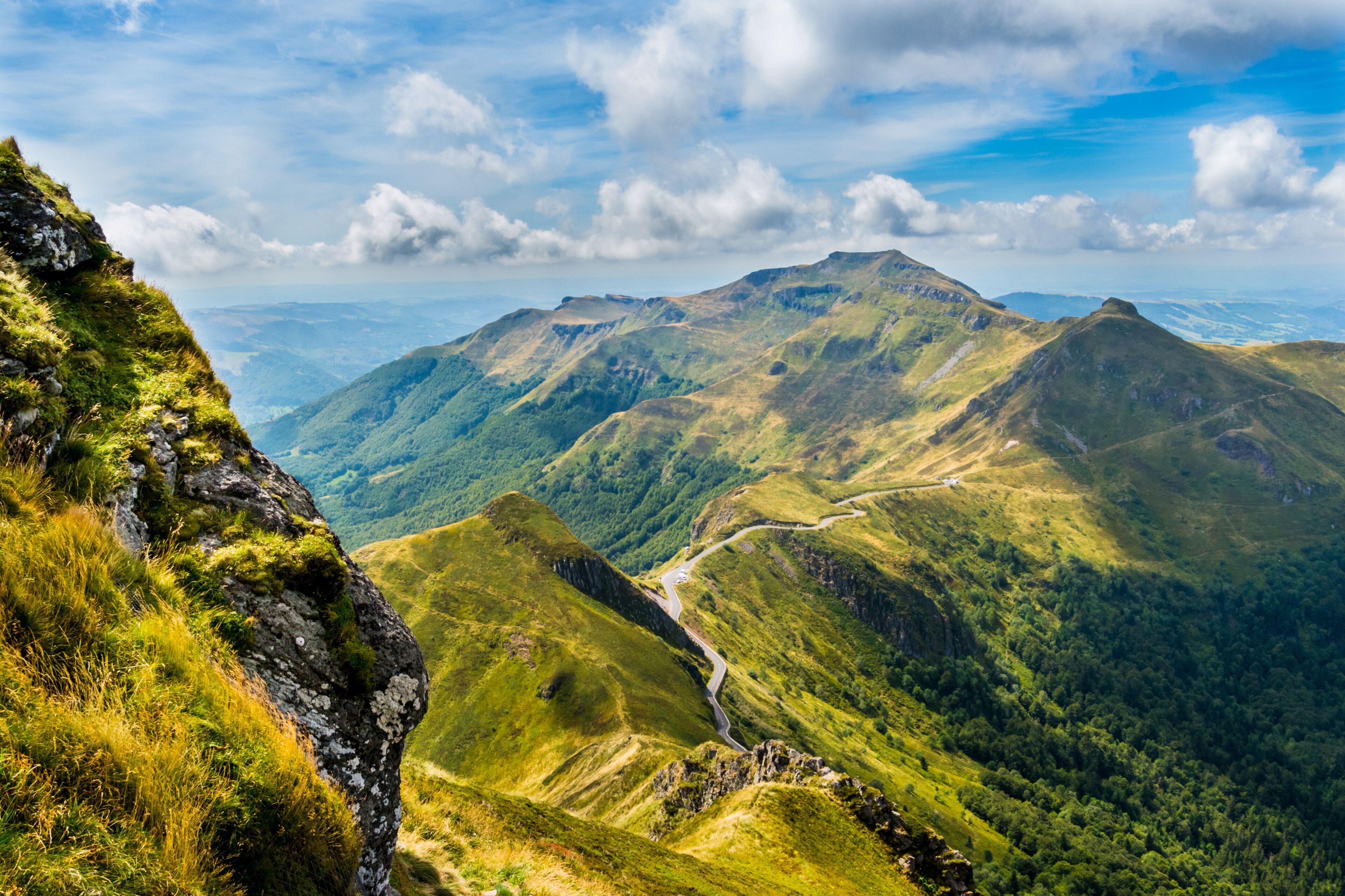 A mountainous landscape with a winding road, lush greenery, and distant hills under a partly cloudy sky.