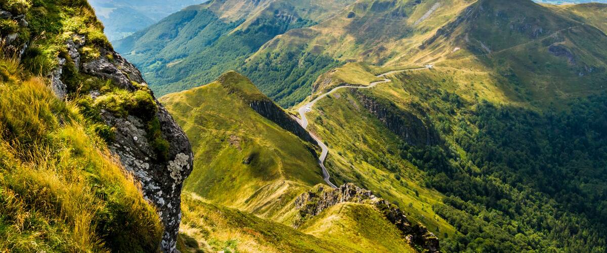 A mountainous landscape with a winding road, lush greenery, and distant hills under a partly cloudy sky.
