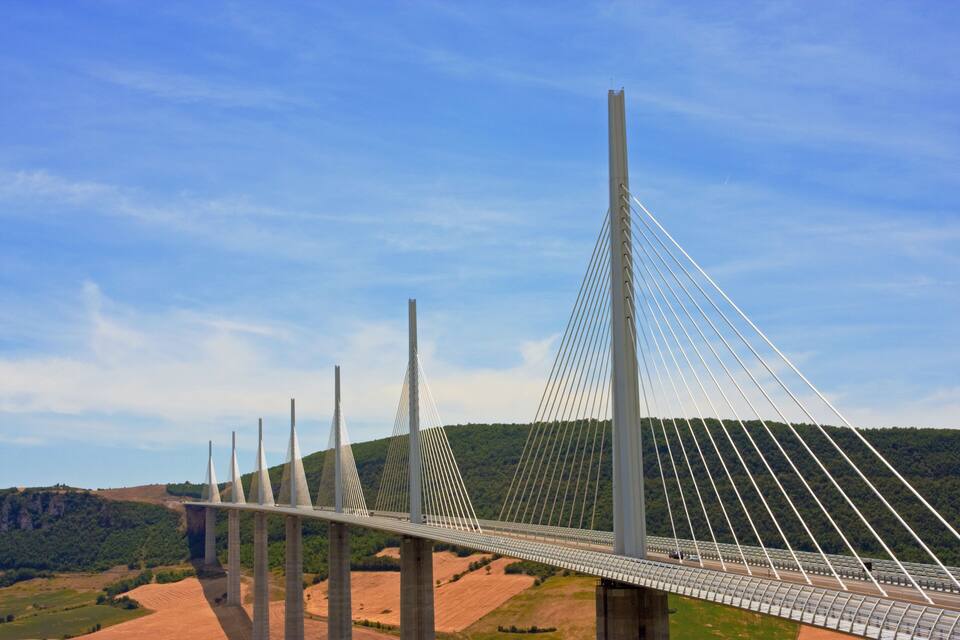 A cable-stayed bridge with multiple towers, set against a backdrop of green hills and a clear blue sky.