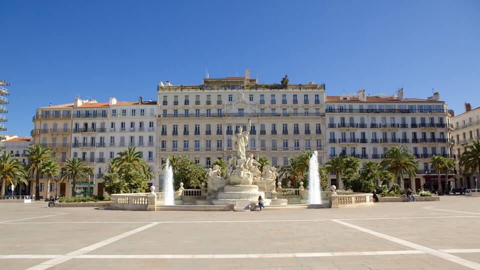 A plaza with a large fountain, palm trees, and historic buildings in the background.