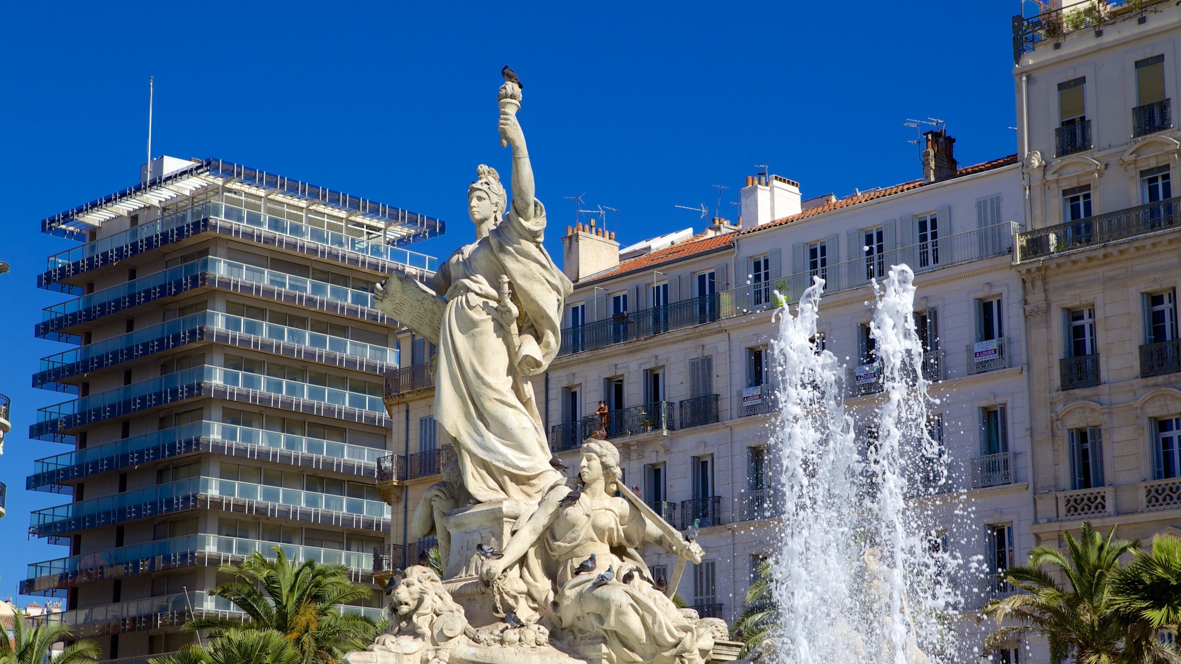 A statue of a woman holding a torch with a lion at her feet, in front of a building with multiple balconies and a fountain.