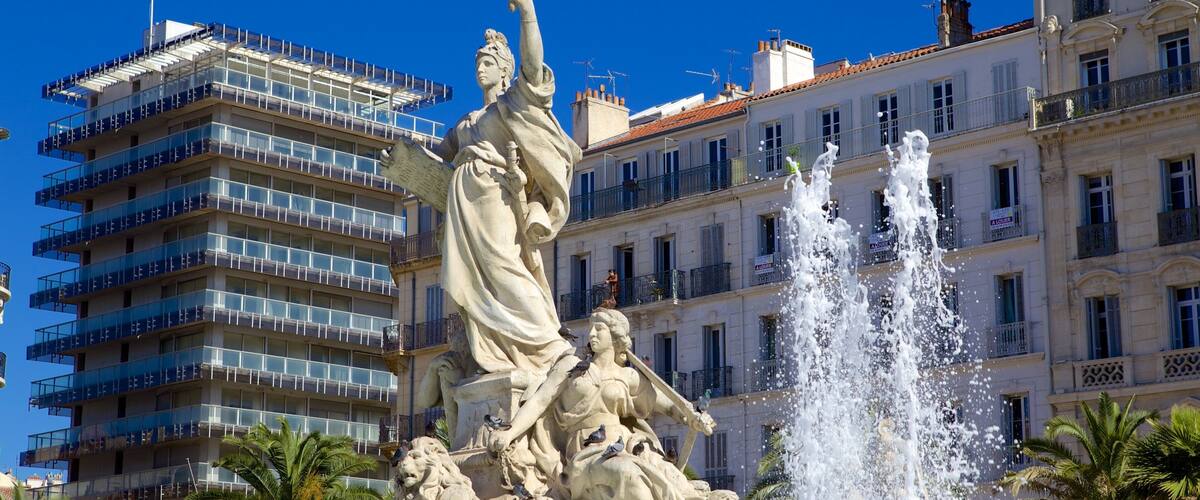 A statue of a woman holding a torch with a lion at her feet, in front of a building with multiple balconies and a fountain.