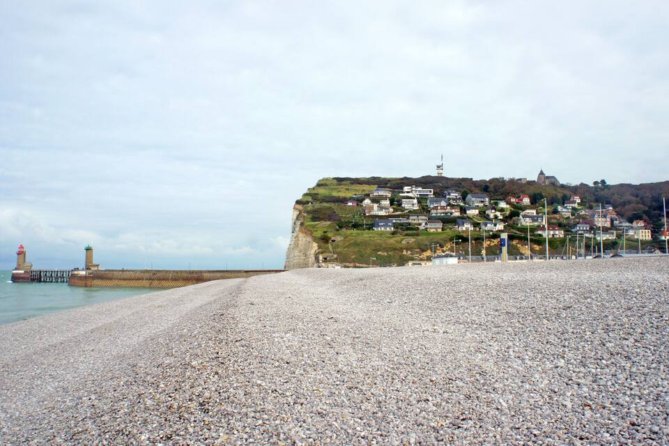 A pebble beach with a lighthouse, houses on a hill, and a church tower.