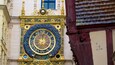 Une horloge détaillée et richement décorée, dotée de chiffres romains et d’un motif solaire, qui est fixée sur la façade d’un bâtiment présentant des éléments d’architecture classique.