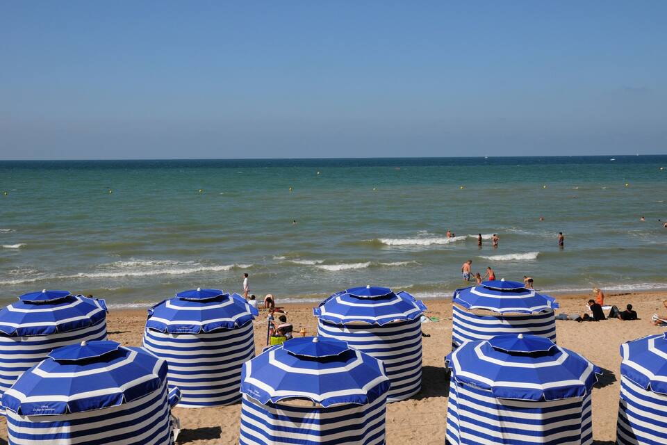 A beach with striped blue and white umbrellas and people swimming in the water.