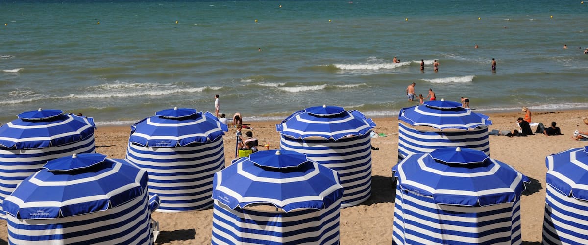 A beach with striped blue and white umbrellas and people swimming in the water.