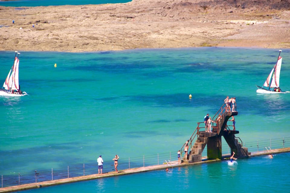 Een kustlandschap met helder turquoise water, een betonnen pier met mensen erop, en twee zeilboten.