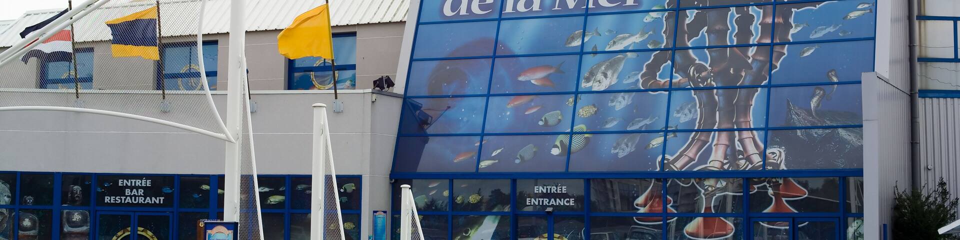 A building with a large mural of an octopus and the text "Mystères de la Mer". There are flags and a sign for an entrance bar and restaurant.