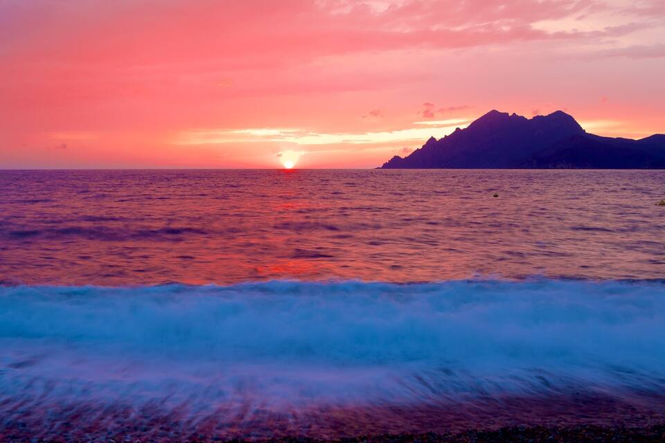 A sunset over the ocean with waves and a mountain in the distance.