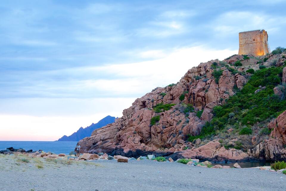 A coastal landscape with a rocky hill, a tower, and a distant mountain range.