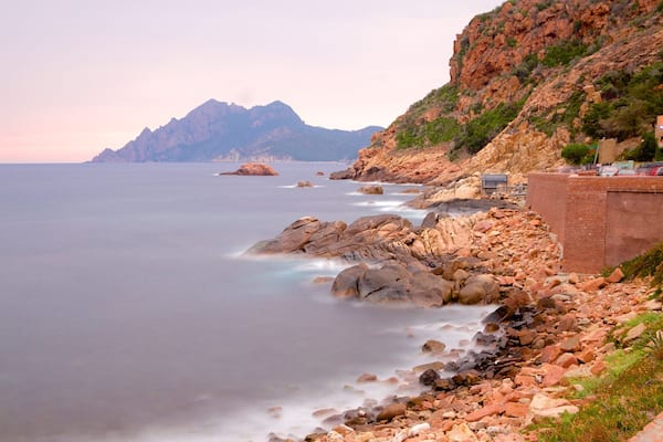 Un paysage côtier caractérisé par un terrain rocheux, une mer calme et une chaîne de montagnes lointaine.