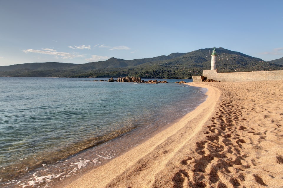 A sandy beach with footprints, a lighthouse, and a mountain in the background.