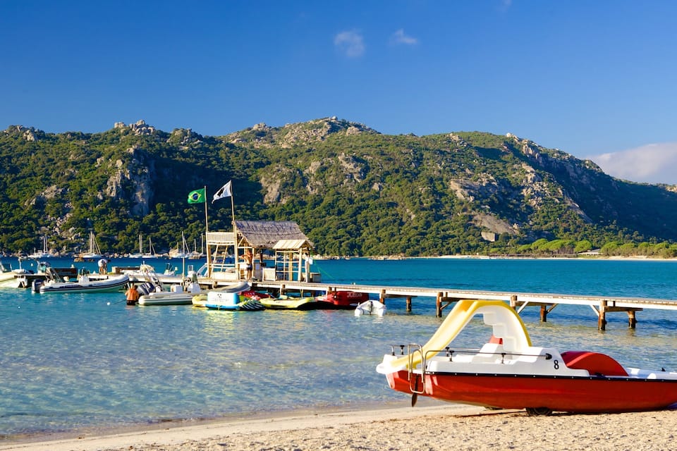 Un paysage côtier avec des bateaux amarrés à un quai, une plage et une chaîne de montagnes en arrière-plan.
