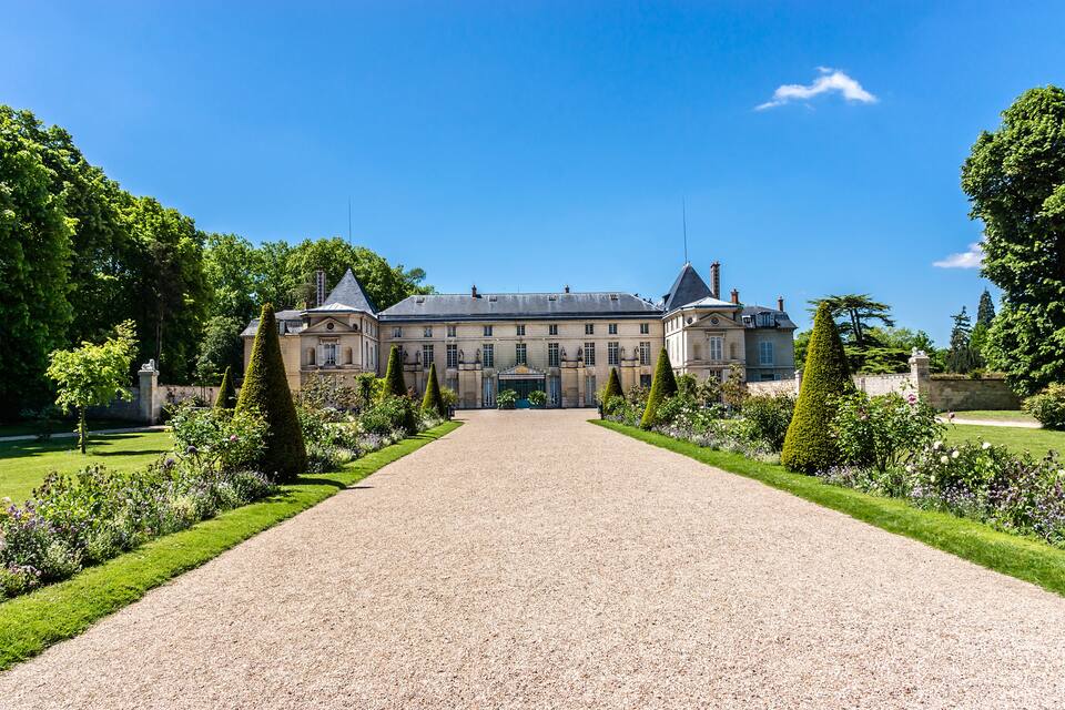 Un edificio grande e histórico con fachada simétrica, rodeado de jardines cuidados y un camino de grava.