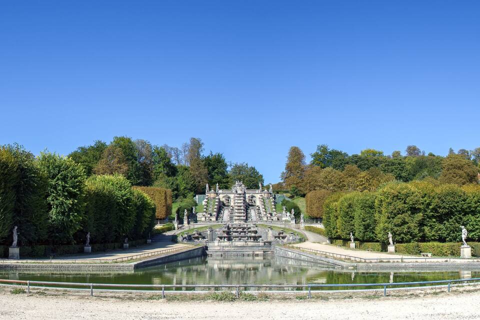 Un'ampia e decorata fontana con statue in un parco alberato, con un edificio sullo sfondo.