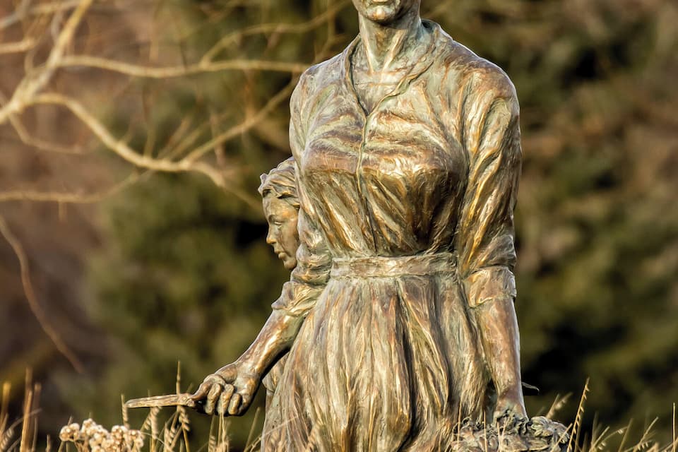 Una estatua de bronce de una mujer sosteniendo a un niño en un campo con césped y árboles al fondo.