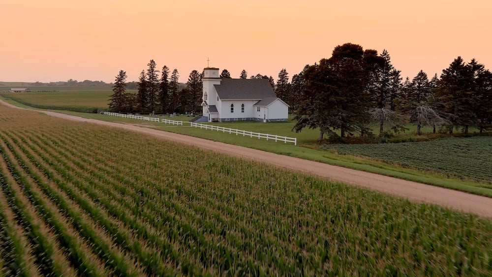 A white church with a steeple surrounded by a white fence, set in a rural landscape with fields and trees.