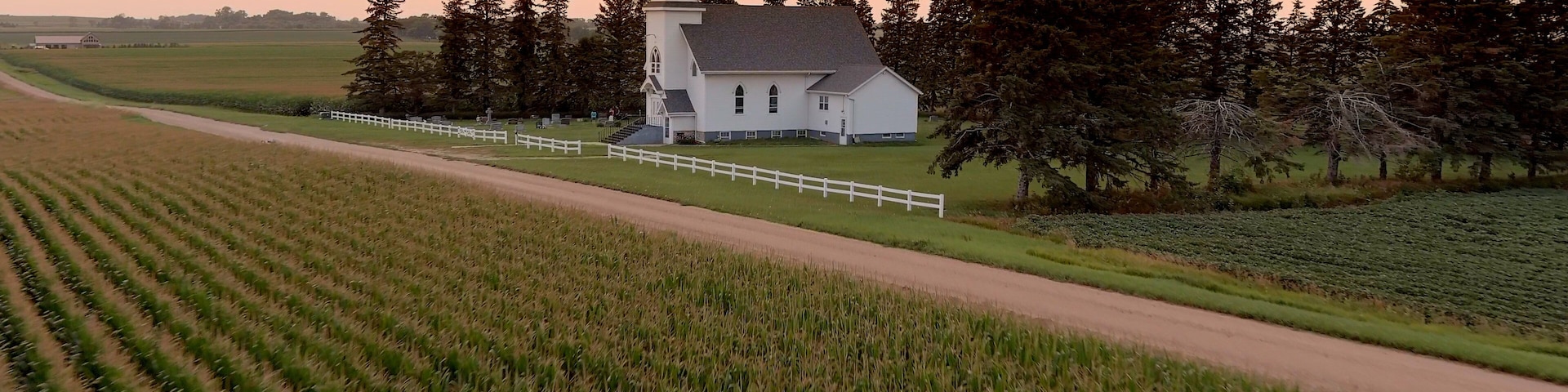 A white church with a steeple surrounded by a white fence, set in a rural landscape with fields and trees.