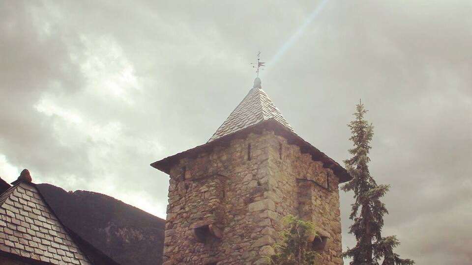 A stone tower with a pointed roof and a weather vane, surrounded by trees and mountains.