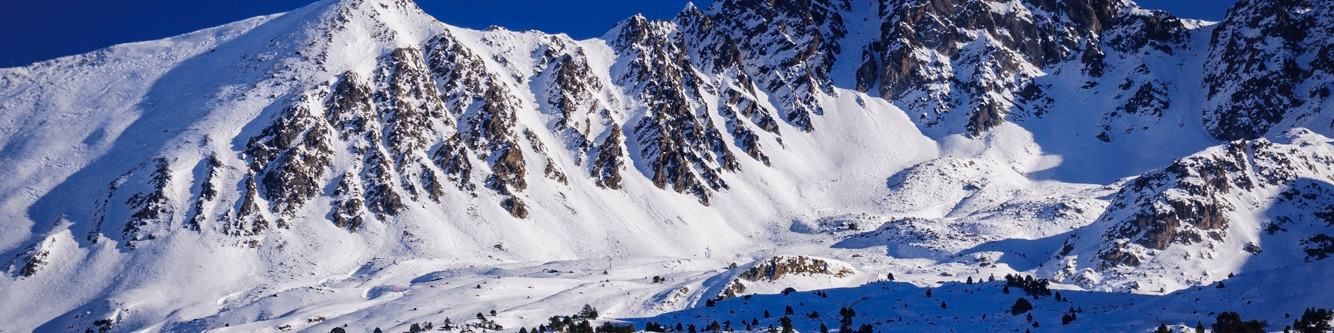 Cordilheira nevada com picos acidentados e árvores dispersas.