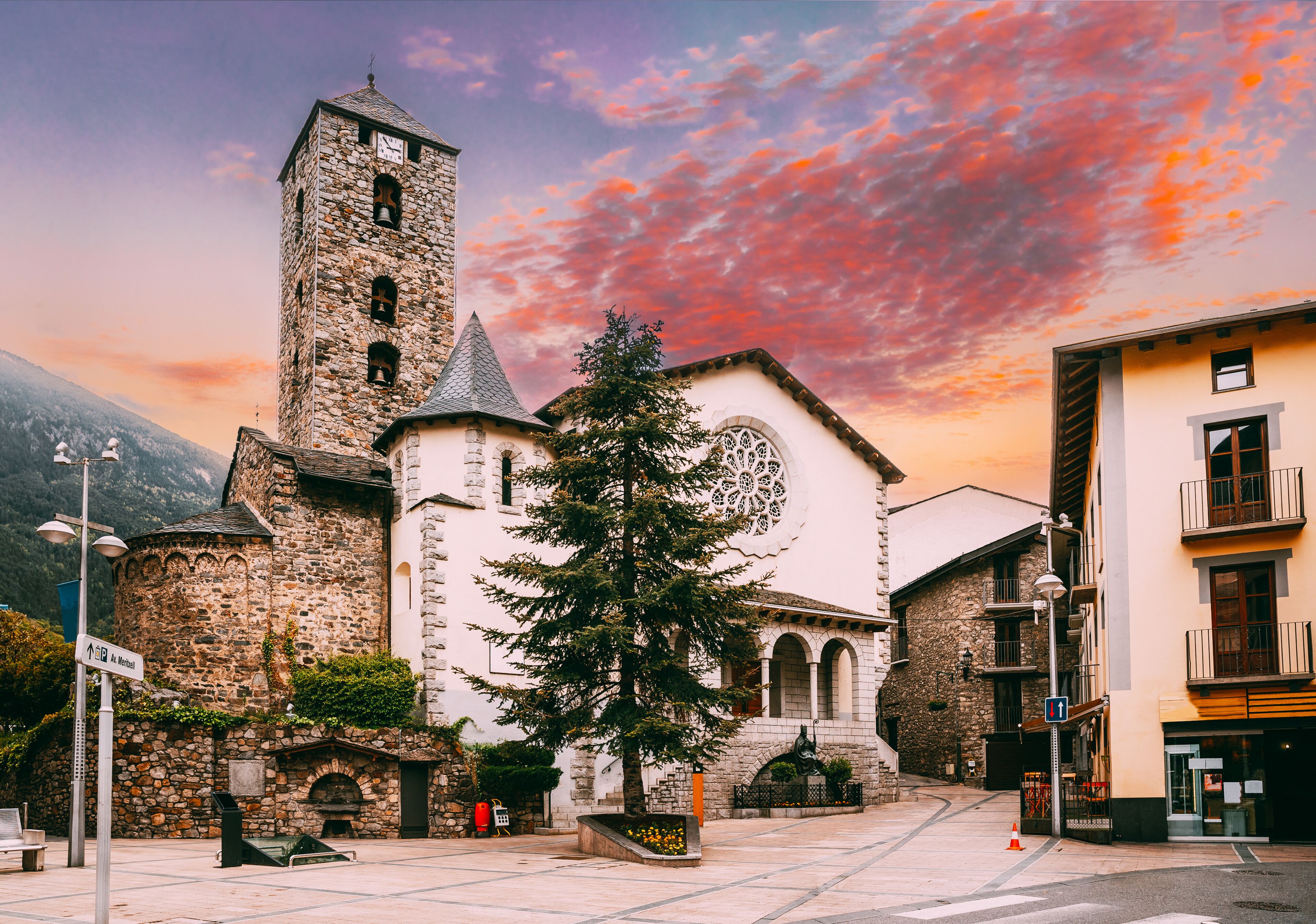 A historic stone church with a bell tower, a large tree, and a mountain backdrop.