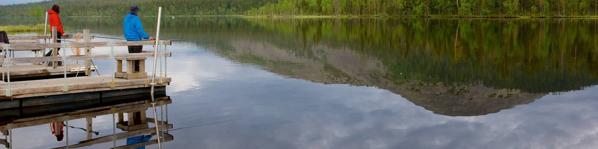 A calm lake with a wooden dock, two people fishing, and a forested hill in the background.