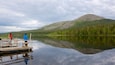 A calm lake with a wooden dock, two people fishing, and a forested hill in the background.