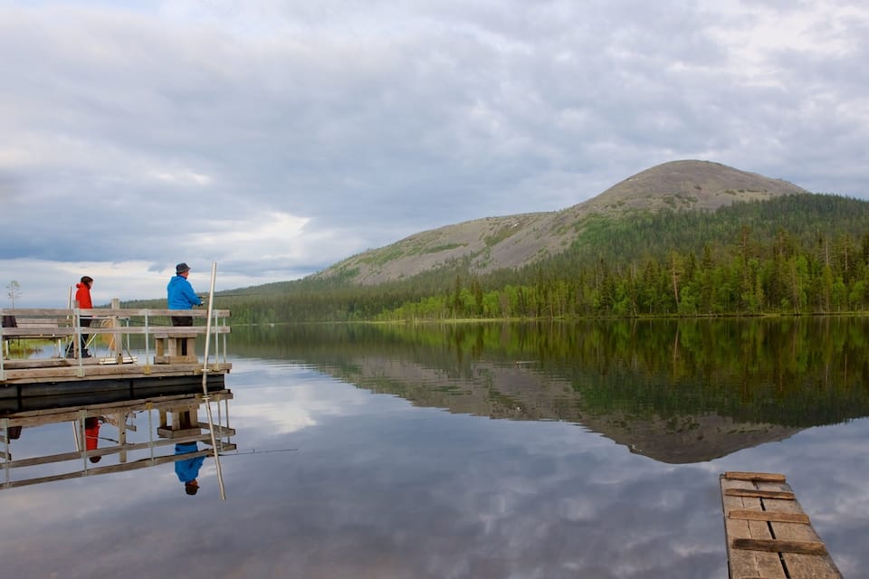 Un lac paisible avec un quai en bois, une personne qui pêche et, au loin, une montagne couverte de forêts.
