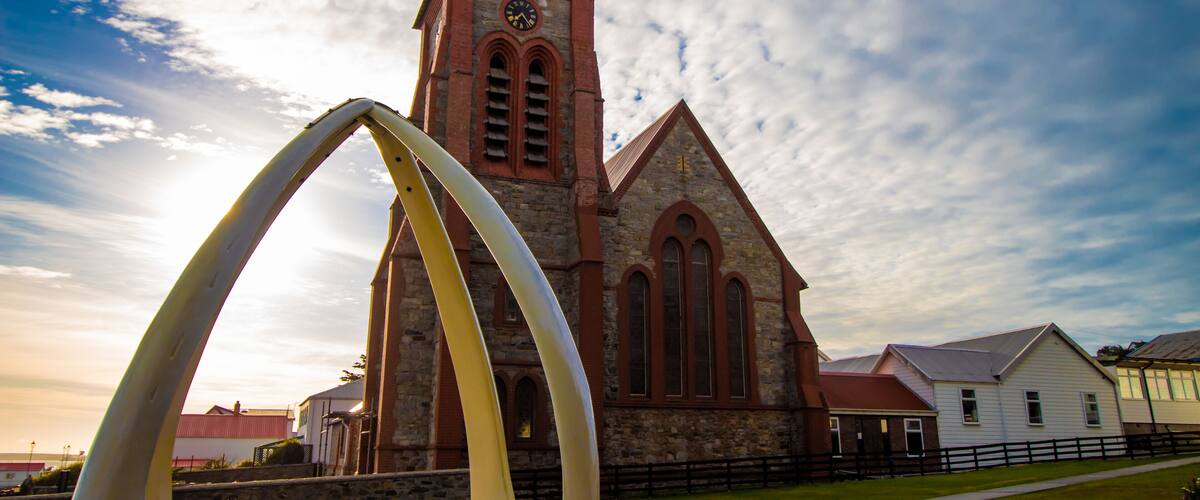 A church with a clock tower, a large arch sculpture, and a white building in the foreground.