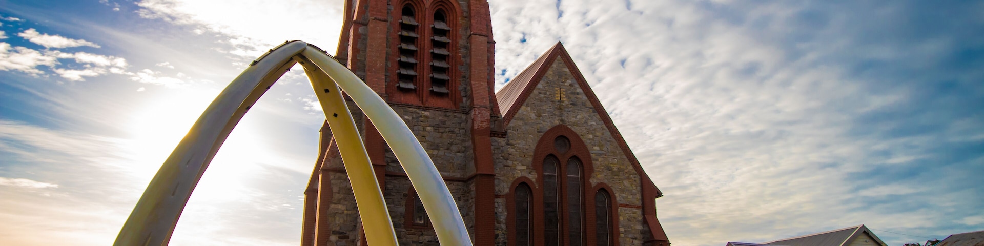 A church with a clock tower, a large arch sculpture, and a white building in the foreground.