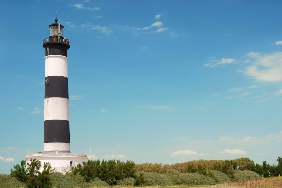 A lighthouse with alternating black and white stripes, surrounded by greenery and a clear blue sky.