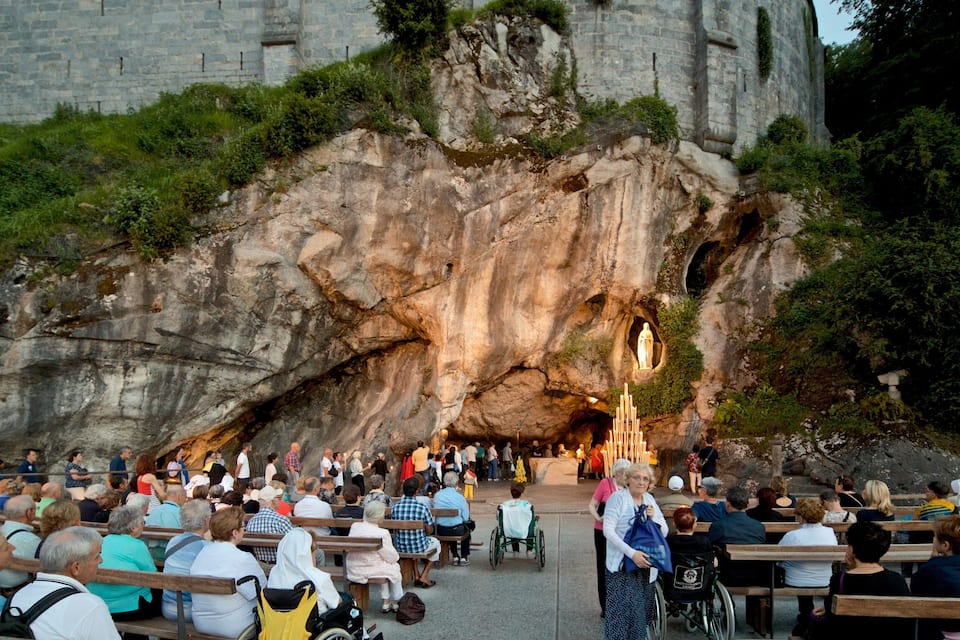 Uma multidão de pessoas reunida em frente a uma grande formação rochosa com uma estátua dentro de uma abertura semelhante a uma caverna.