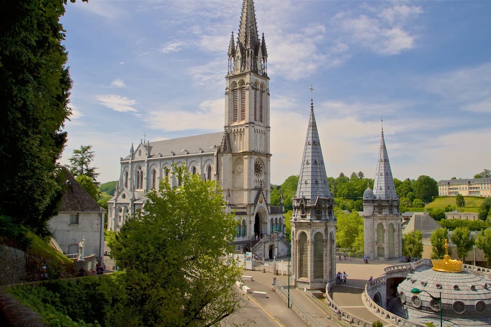 Uma catedral histórica com duas torres gêmeas e uma torre central imponente, cercada por vegetação exuberante e uma praça circular com um monumento em forma de coroa dourada.