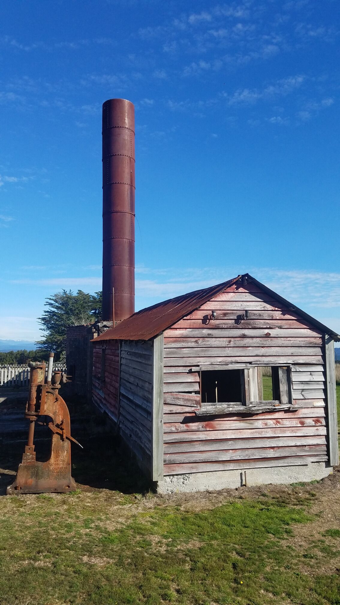 A rusted metal structure with a tall chimney, a wooden building with a gabled roof, and a grassy field.