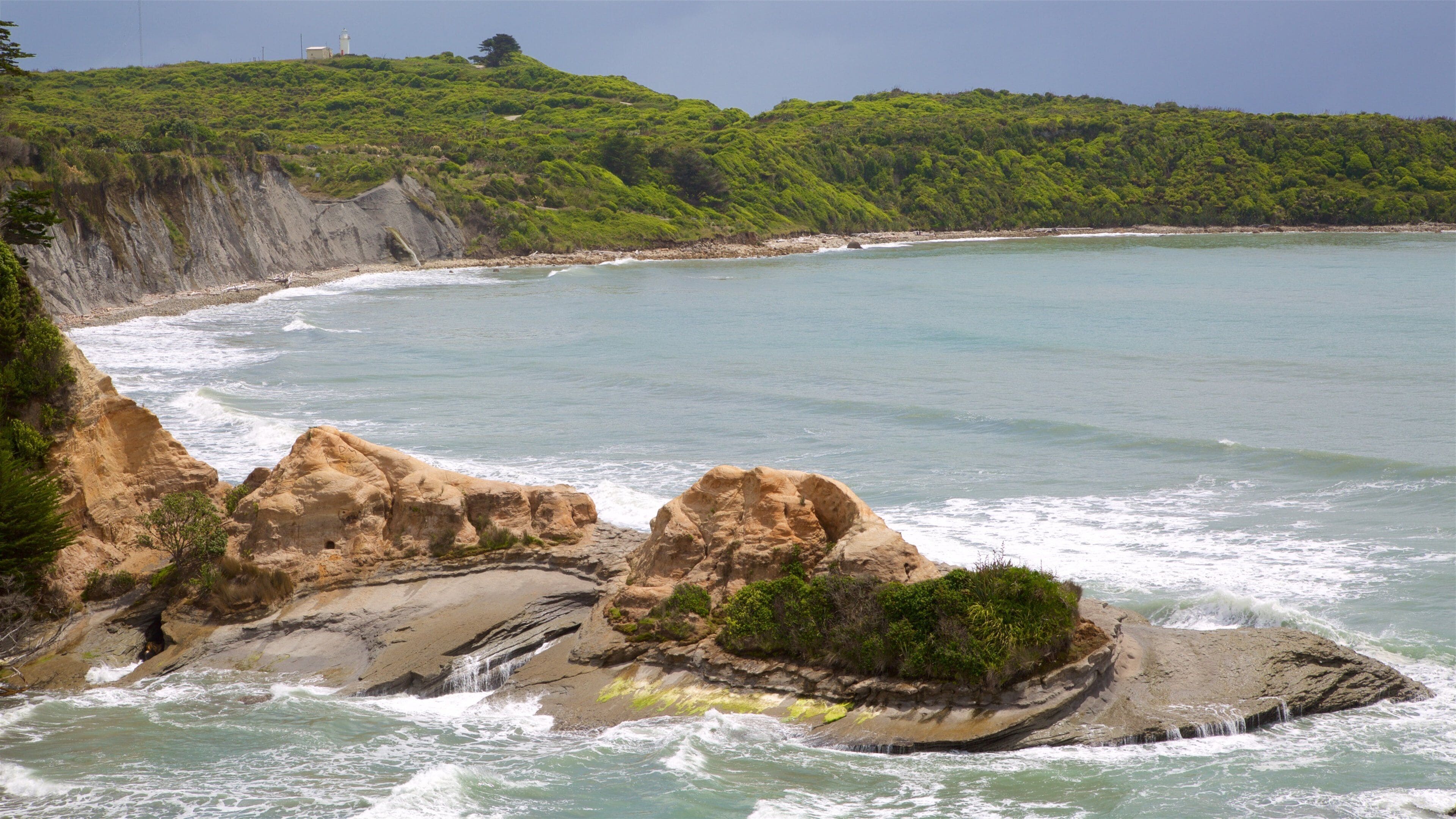 A coastal landscape with rocky formations, a beach, and a lighthouse in the distance.