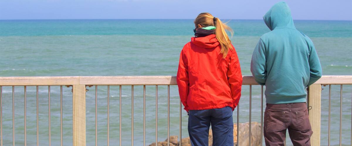 Two people standing on a wooden railing overlooking a rocky shoreline with calm sea water.