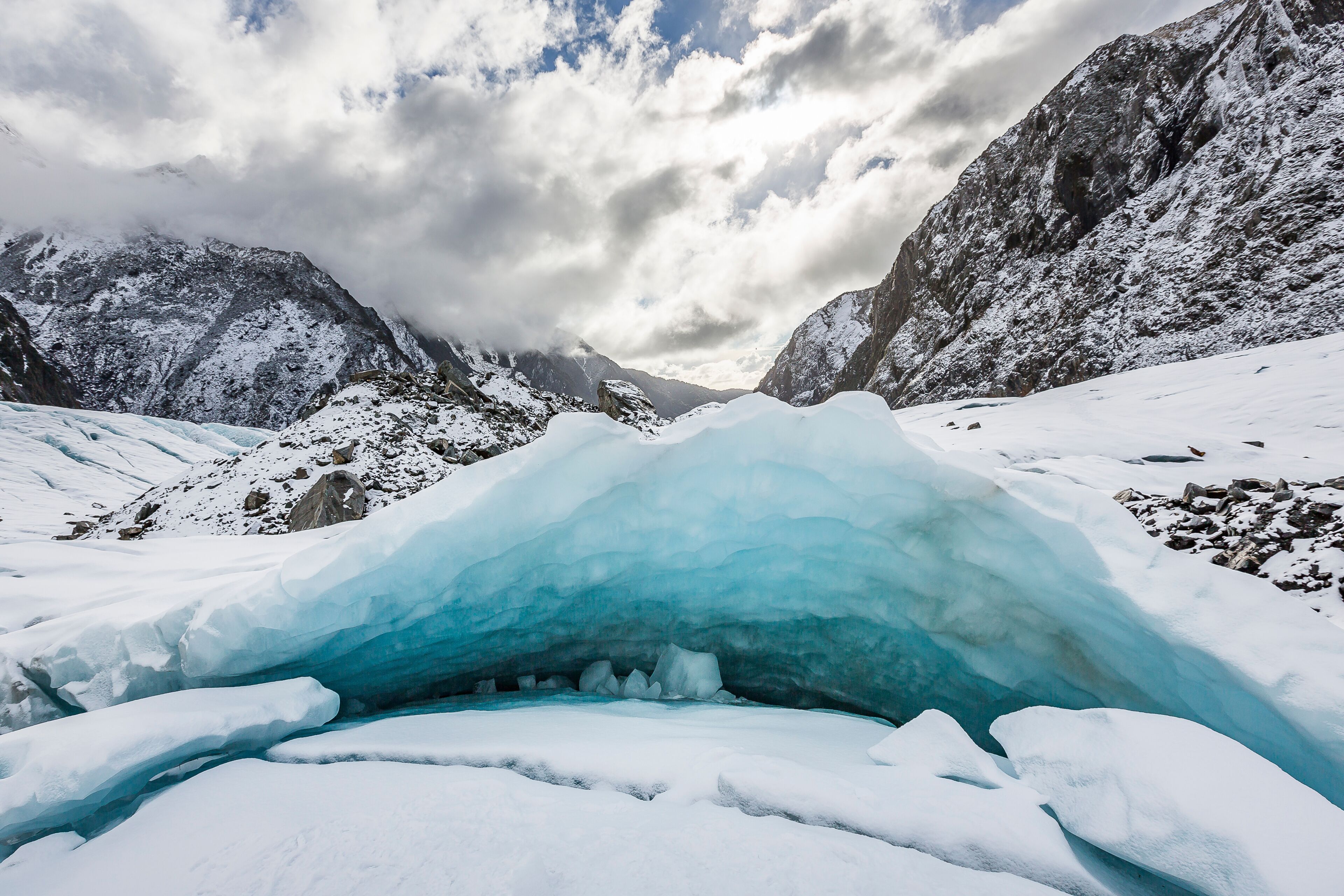 A glacier with a crevasse, snow-covered mountains, and a partly cloudy sky.