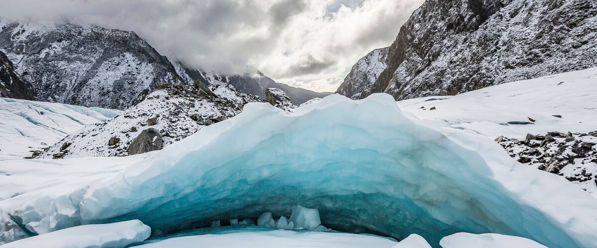 A glacier with a crevasse, snow-covered mountains, and a partly cloudy sky.