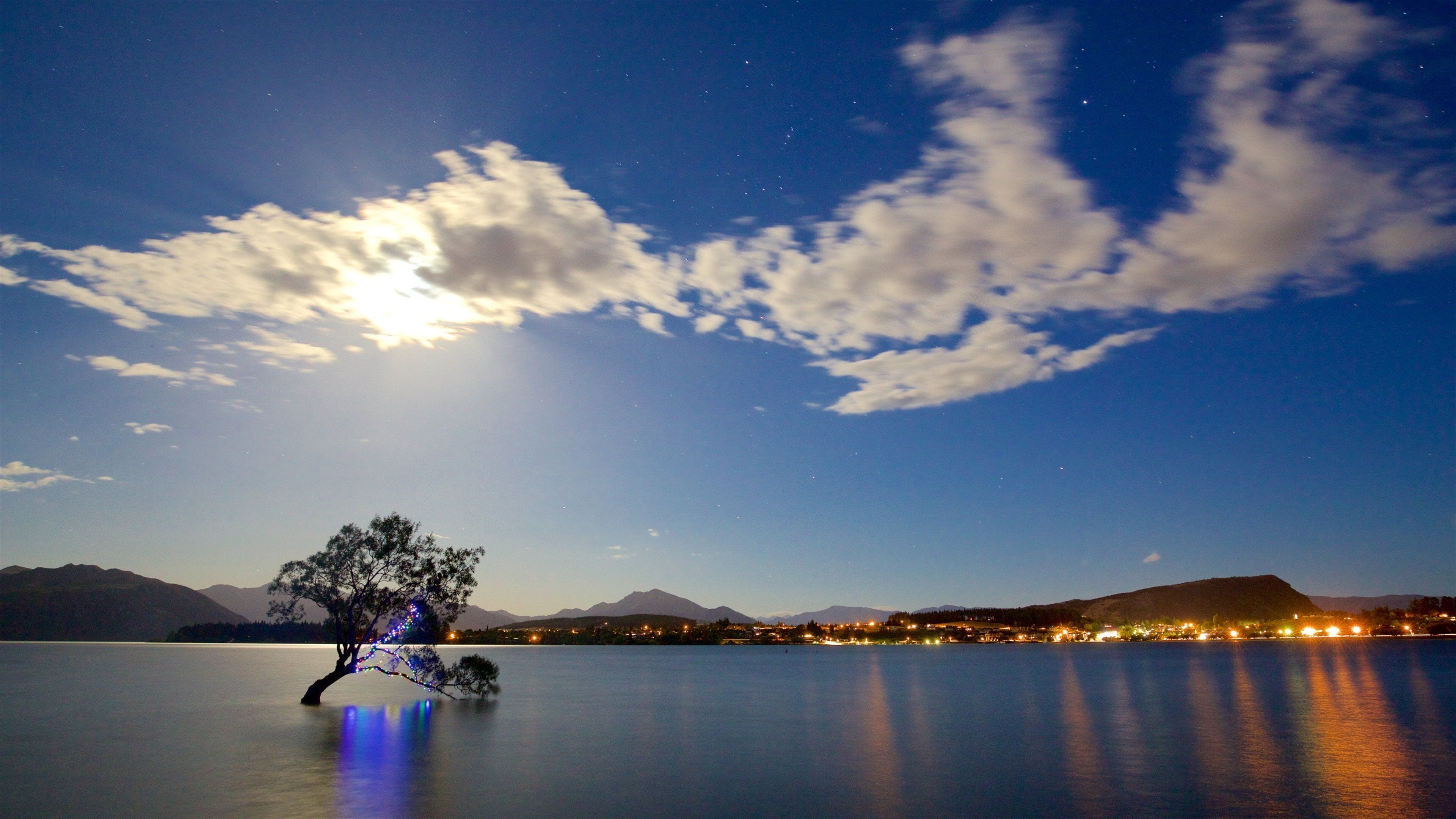 A night scene with a lake, a tree with colorful lights, mountains, and a city skyline with lights reflecting on the water.