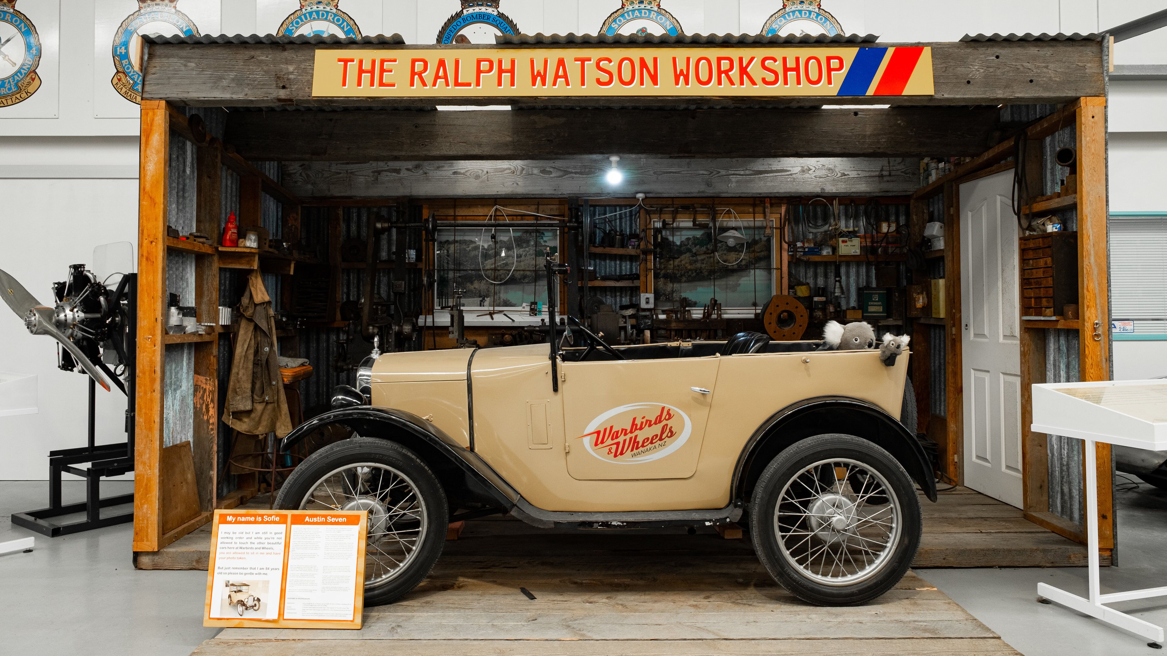 A vintage car in a workshop with a sign reading "The Ralph Watson Workshop".