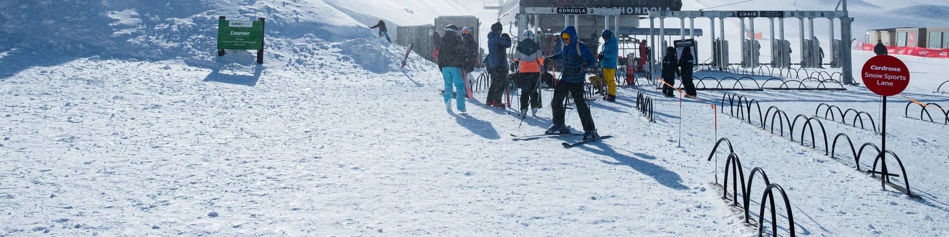 Pista de esquí nevada con gente esquiando y de pie, un letrero que indica 'Adulto / Principiante' y un letrero de 'Cardrona Snow Sports Lane'.