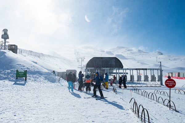 A snowy ski slope with people skiing and standing, a sign for 'Adult / Learner', and a 'Cardrona Snow Sports Lane' sign.