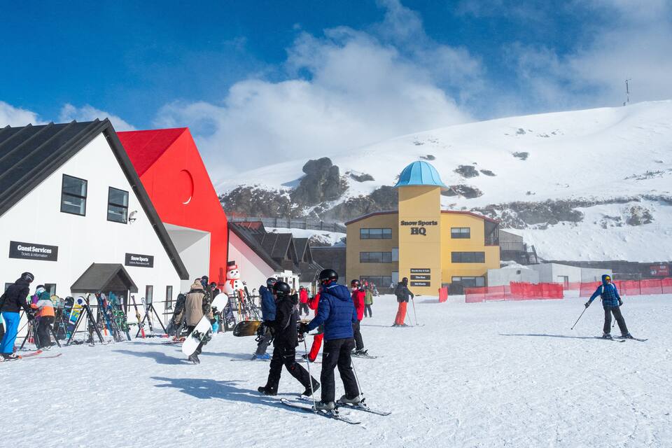 Et snøklædt skisportssted med en rød og hvid bygning med skiltet "Guest Services" og en gul bygning med skiltene "Snow Sports HQ".