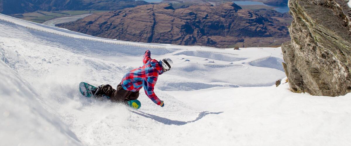 A snowboarder in a red plaid jacket and helmet, descending a snowy slope with a backdrop of rugged mountains and a lake.