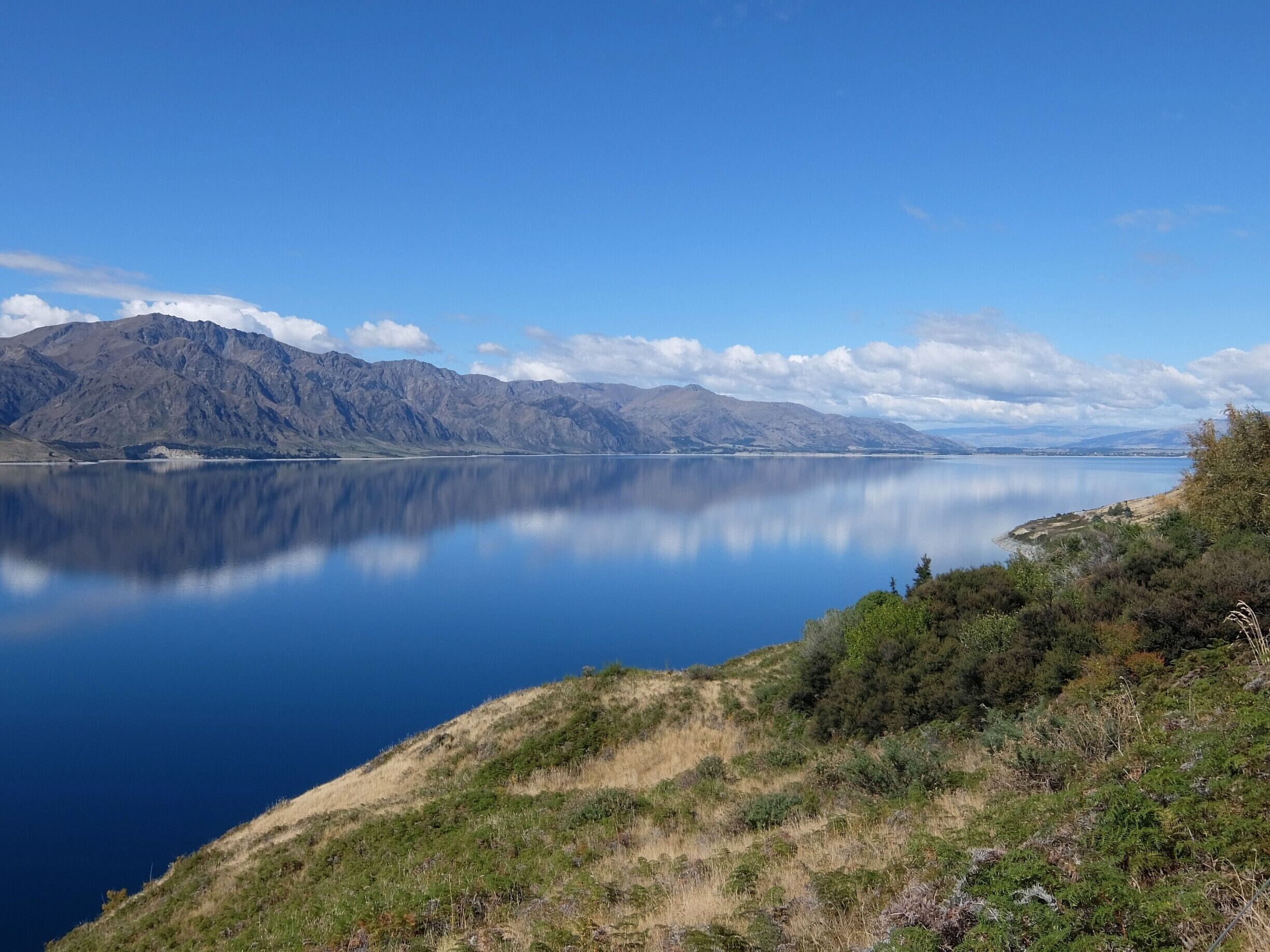 A calm lake with mountains in the background and a grassy hill in the foreground.