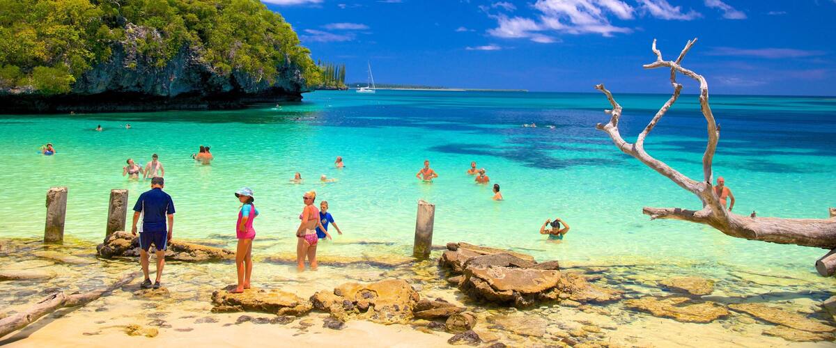 A tropical beach with clear turquoise water, people swimming and standing on rocks, and a lush green island in the background.