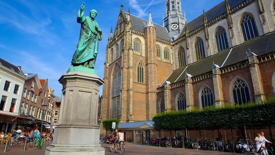 Eine Statue auf einem Platz, eine historische Kirche mit einem Glockenturm und eine belebte Straße mit Fußgängern und Fahrrädern.