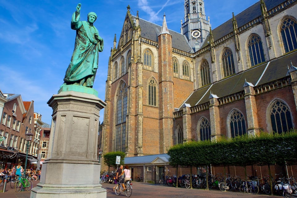 Una estatua en una plaza, una iglesia histórica con una torre de campanario y una calle concurrida con peatones y bicicletas.