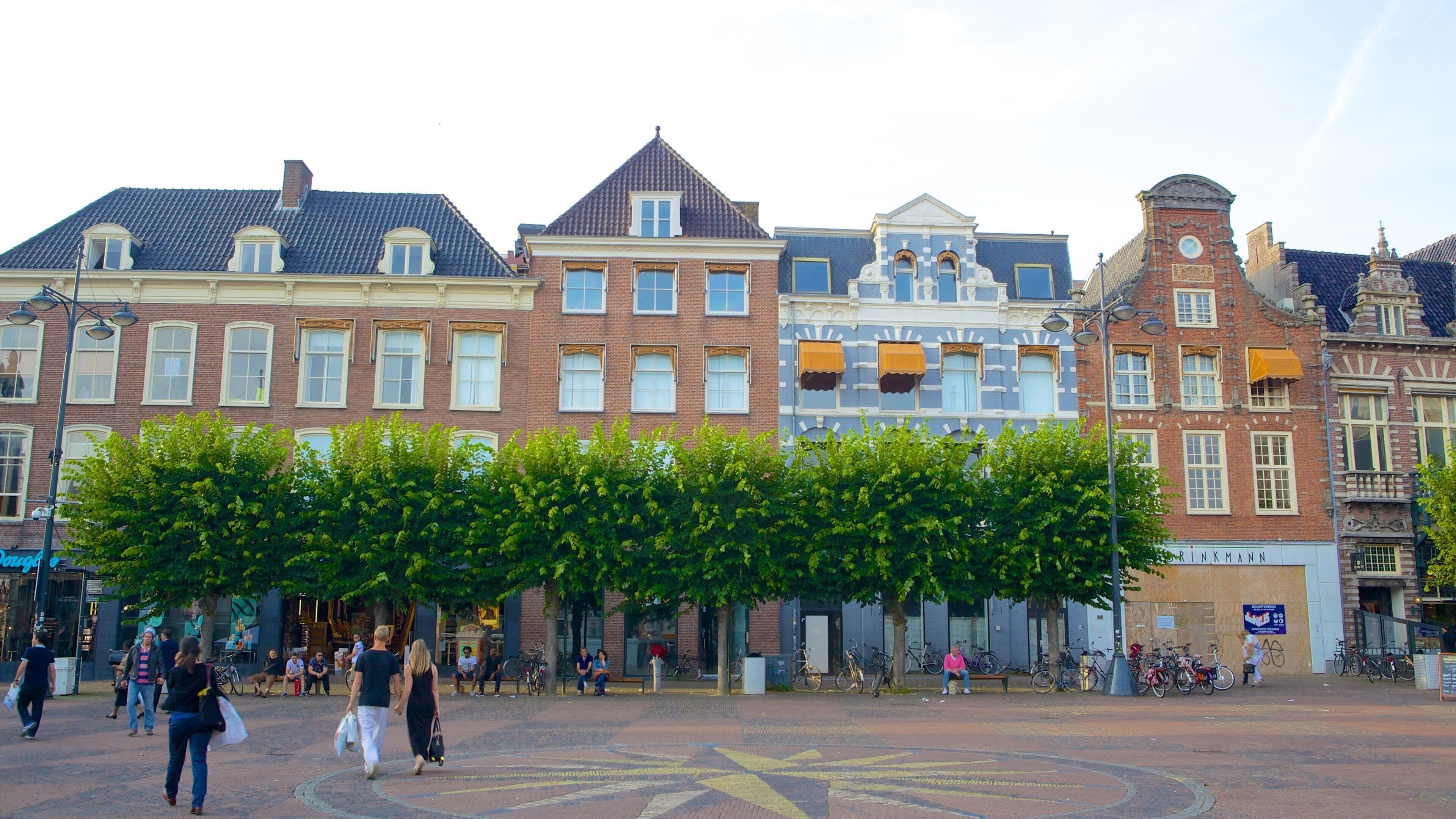 Uma rua histórica europeia com arquitetura tradicional, uma praça central arborizada e pessoas caminhando.