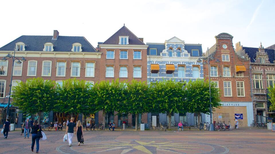 Eine historische europäische Straße mit traditioneller Architektur, ein zentraler, von Bäumen gesäumter Platz und Passanten.